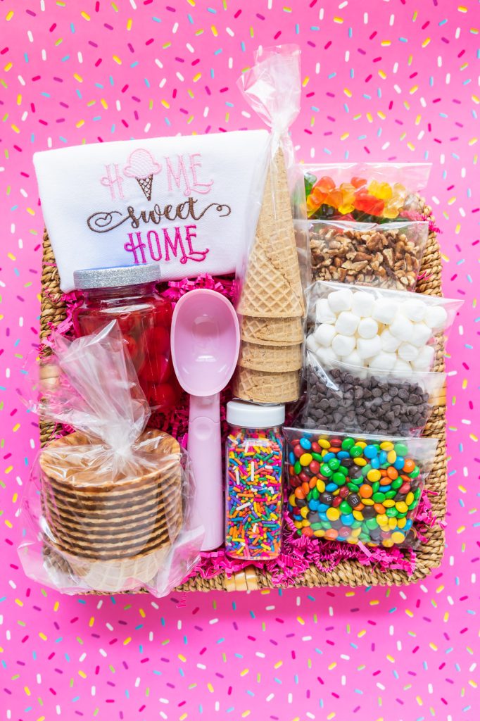 basket filled with wrapped ice cream toppings, jar of cherries, ice cream scoop, waffle bowls, and handmade towel